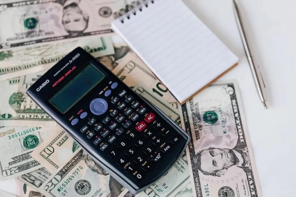 Calculator, notepad, pen, and scattered US dollar bills on a table.