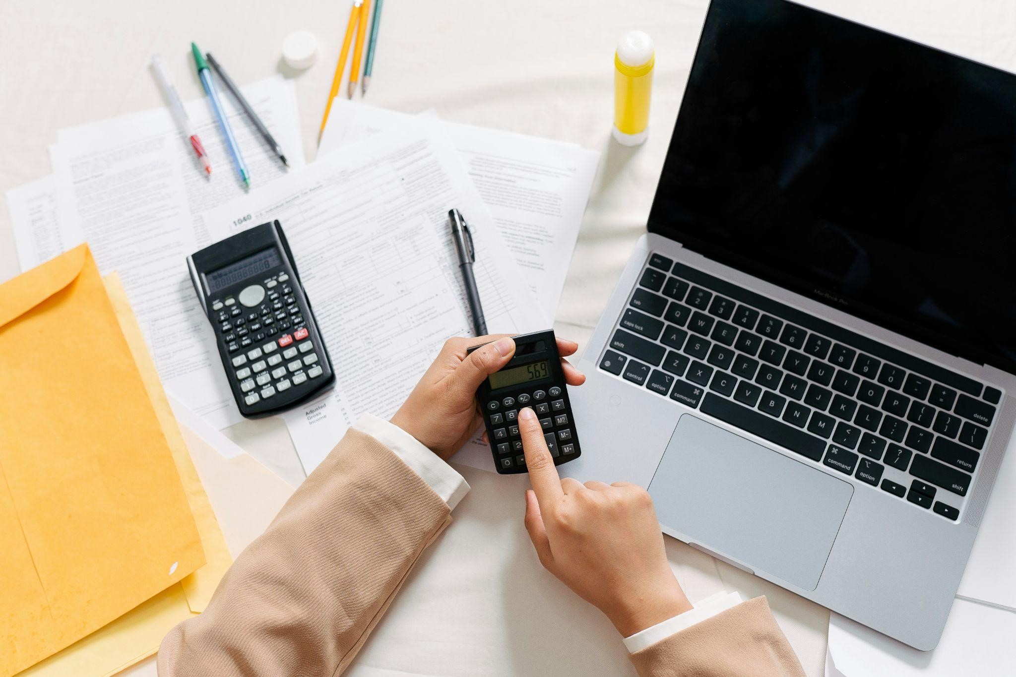 Person using calculator at desk with laptop and documents.