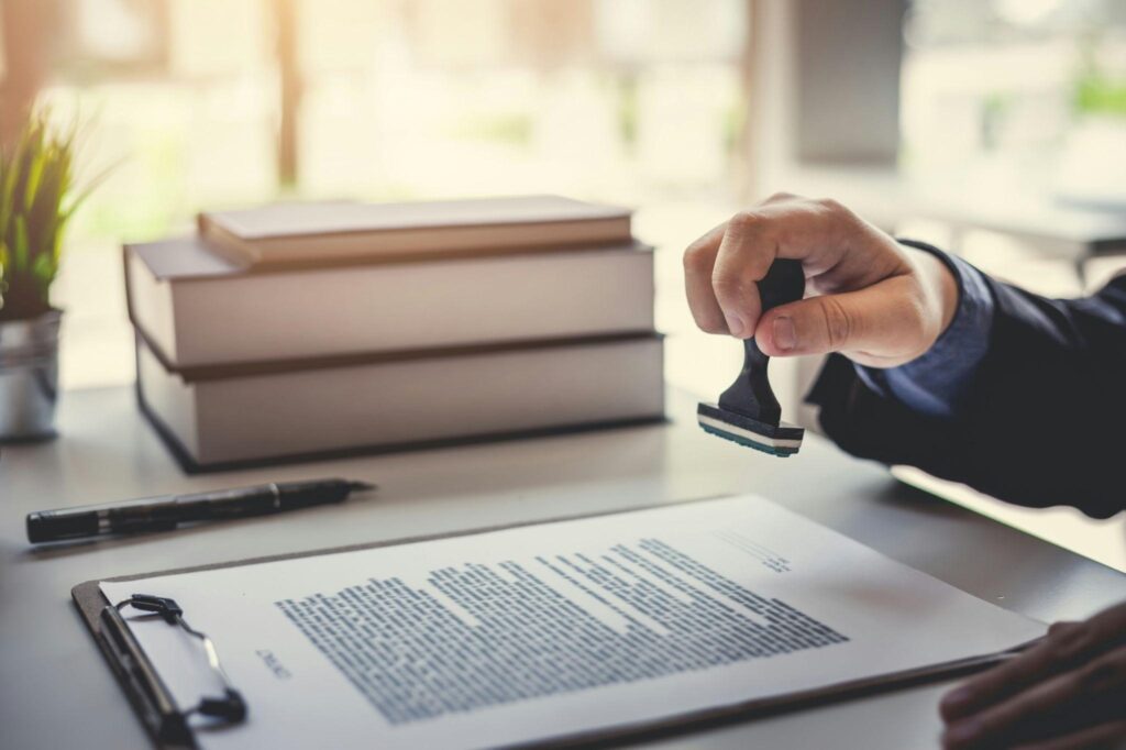Hand stamping a document on a clipboard, with books in the background.