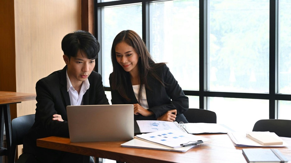 Two individuals watching a laptop with papers and files on a table.
