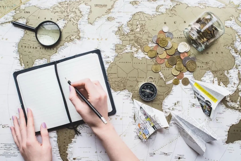 A journal, magnifying glass, compass, coins, and paper planes on top of a world map