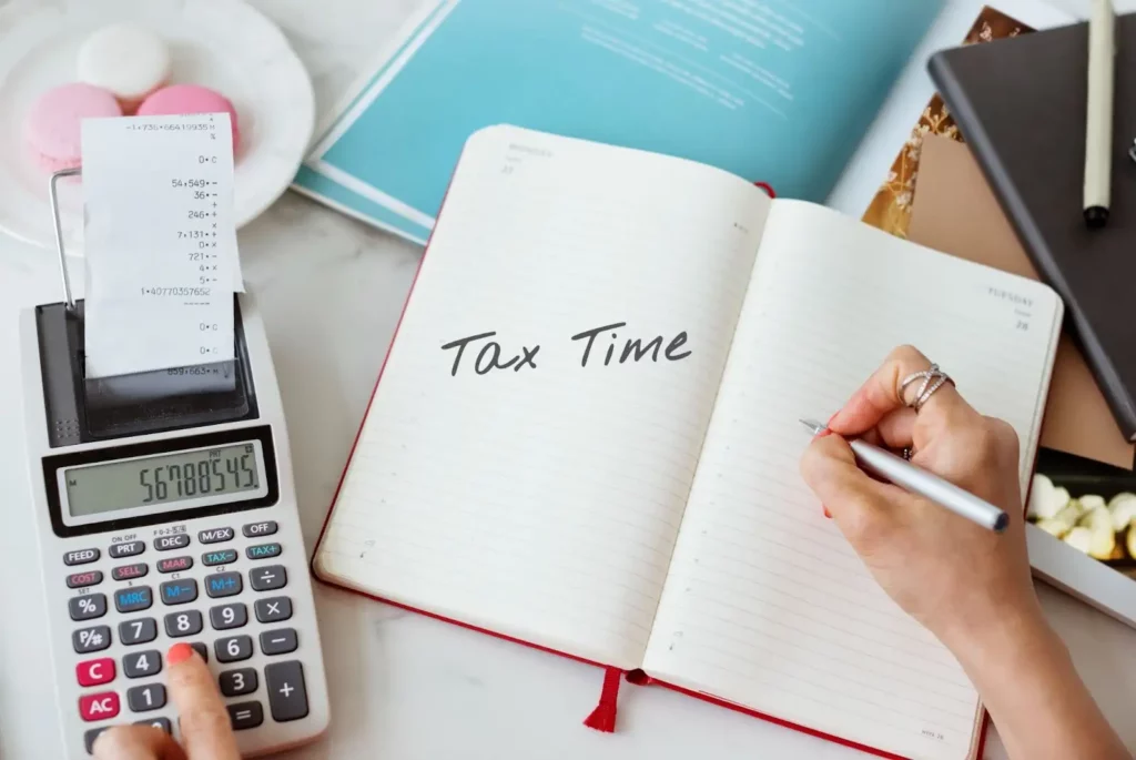 A person using a calculator, a notebook, and a pen to compute tax