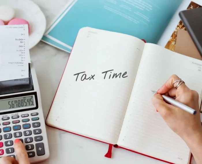 A person using a calculator, a notebook, and a pen to compute tax